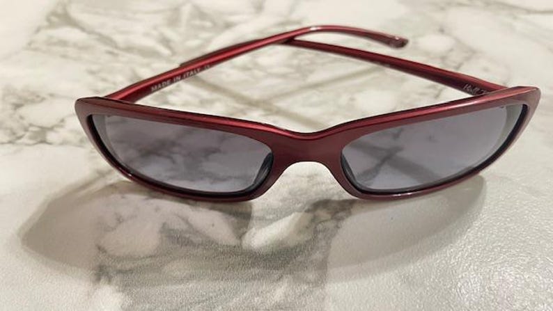 May include: Red-framed sunglasses with gray lenses. The sunglasses have a rectangular shape and a glossy finish. The image shows the sunglasses on a marble-patterned surface. The text "MADE IN ITALY" is visible on the frame.