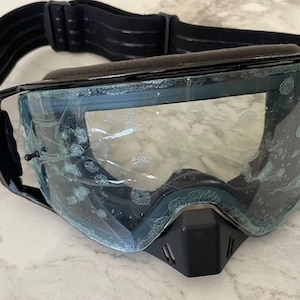 Può includere: Occhiali protettivi neri con lente trasparente coperta da una pellicola protettiva in plastica, montatura nera e cinturino elastico nero regolabile. Gli occhiali includono una protezione nasale con piccole fessure di ventilazione frontali.