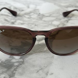 May include: A pair of sunglasses with a dark purple frame and brown lenses. The sunglasses have gold-colored arms and the Ray-Ban logo is visible on the left lens. The glasses are set against a white and gray marble background.