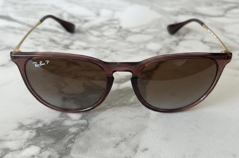 May include: A pair of sunglasses with a dark purple frame and brown lenses. The sunglasses have gold-colored arms and the Ray-Ban logo is visible on the left lens. The glasses are set against a white and gray marble background.