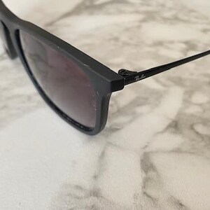 Puede incluir: Gafas de sol negras con lentes oscuras. Las gafas tienen una montura rectangular y finas patillas negras. El logotipo de Ray-Ban es visible en la patilla. Las gafas descansan sobre una superficie de m&aacute;rmol blanco y gris.