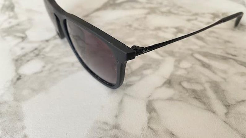 Puede incluir: Gafas de sol negras con lentes oscuras. Las gafas tienen una montura rectangular y finas patillas negras. El logotipo de Ray-Ban es visible en la patilla. Las gafas descansan sobre una superficie de m&aacute;rmol blanco y gris.