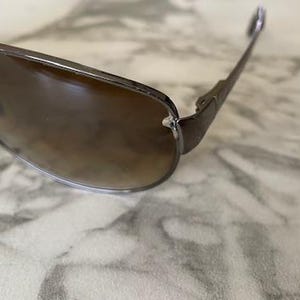 May include: Close-up of sunglasses with a brown gradient lens and a dark metal frame. The sunglasses have a classic aviator style with a sleek, modern design. The lenses are slightly tinted, offering a stylish look.