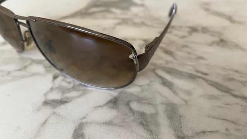 May include: Close-up of sunglasses with a brown gradient lens and a dark metal frame. The sunglasses have a classic aviator style with a sleek, modern design. The lenses are slightly tinted, offering a stylish look.