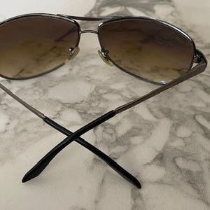 May include: Aviator-style sunglasses with a silver metal frame and gradient brown lenses. The arms are thin and extend to black tips, crossing at the ends. The sunglasses are designed for sun protection and fashion.