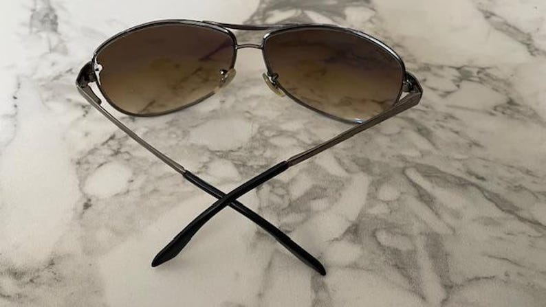 May include: Aviator-style sunglasses with a silver metal frame and gradient brown lenses. The arms are thin and extend to black tips, crossing at the ends. The sunglasses are designed for sun protection and fashion.