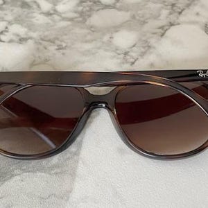 May include: Brown sunglasses with a tortoiseshell pattern. The sunglasses have a classic aviator shape with dark lenses and the Ray-Ban logo on the temples. The frame is a dark brown color.