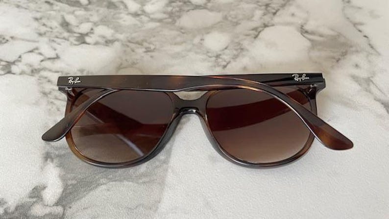 May include: Brown sunglasses with a tortoiseshell pattern. The sunglasses have a classic aviator shape with dark lenses and the Ray-Ban logo on the temples. The frame is a dark brown color.