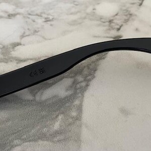 May include: Black sunglasses arm with a red "USA MADE" logo. The arm curves slightly and has a CE mark. The background is a light gray and white marble pattern.