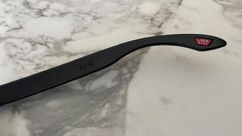May include: Black sunglasses arm with a red "USA MADE" logo. The arm curves slightly and has a CE mark. The background is a light gray and white marble pattern.