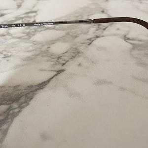 May include: Close-up of sunglasses with a brown tortoiseshell frame and dark lenses. The temples are silver and brown, with the text "Made in Thailand" visible. The sunglasses are on a marble surface.