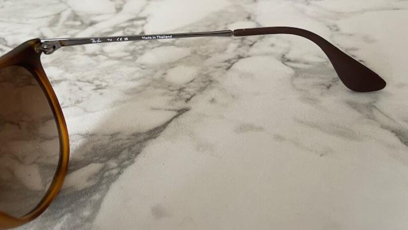 May include: Close-up of sunglasses with a brown tortoiseshell frame and dark lenses. The temples are silver and brown, with the text "Made in Thailand" visible. The sunglasses are on a marble surface.