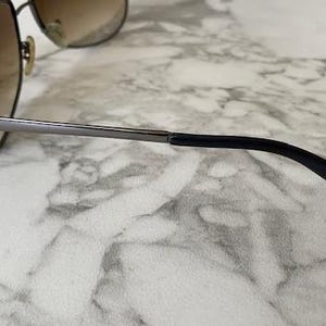 May include: Aviator-style sunglasses with a brown gradient lens and a silver metal frame. The temples are black and curve down towards the ear. The sunglasses are resting on a white and gray marble surface.