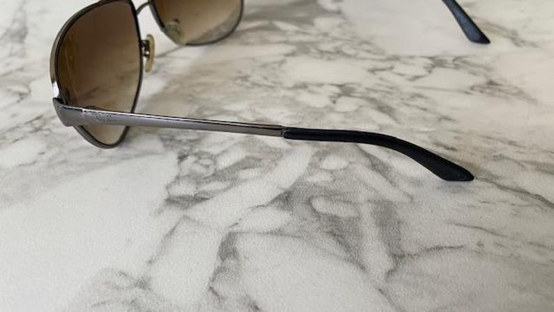 May include: Aviator-style sunglasses with a brown gradient lens and a silver metal frame. The temples are black and curve down towards the ear. The sunglasses are resting on a white and gray marble surface.