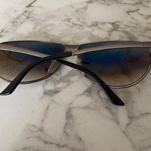 May include: Sunglasses with a silver frame and gradient lenses. The lenses transition from brown to blue. The temples are black and the sunglasses are resting on a marble surface.