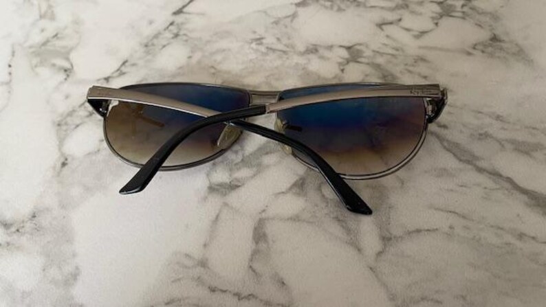 May include: Sunglasses with a silver frame and gradient lenses. The lenses transition from brown to blue. The temples are black and the sunglasses are resting on a marble surface.