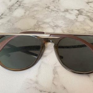 K&ouml;nnte beinhalten: Sonnenbrille mit dunklen Gl&auml;sern und einem braunen Rahmen. Der obere Steg ist silberfarben. Die B&uuml;gel sind braun und erstrecken sich vom Rahmen, um sich um die Ohren zu legen. Die Sonnenbrille liegt auf einer wei&szlig;-grauen Marmoroberfl&auml;che.