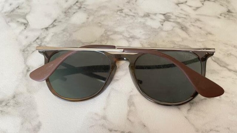 K&ouml;nnte beinhalten: Sonnenbrille mit dunklen Gl&auml;sern und einem braunen Rahmen. Der obere Steg ist silberfarben. Die B&uuml;gel sind braun und erstrecken sich vom Rahmen, um sich um die Ohren zu legen. Die Sonnenbrille liegt auf einer wei&szlig;-grauen Marmoroberfl&auml;che.