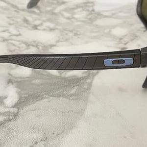 May include: Dark gray sunglasses with purple lenses. The temples have a textured design and a light blue accent with the brand logo. The sunglasses are set against a marble background.