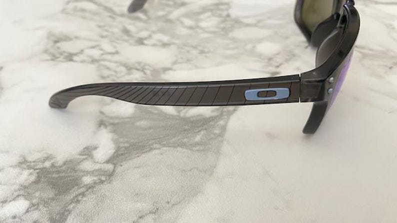 May include: Dark gray sunglasses with purple lenses. The temples have a textured design and a light blue accent with the brand logo. The sunglasses are set against a marble background.