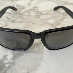 May include: Black sunglasses with a rectangular frame and dark lenses. The sunglasses have a thick frame with small silver accents on the corners. The temples curve inward for a secure fit. The lenses are a dark gray color.