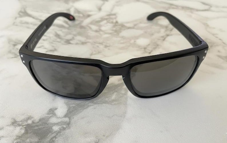 May include: Black sunglasses with a rectangular frame and dark lenses. The sunglasses have a thick frame with small silver accents on the corners. The temples curve inward for a secure fit. The lenses are a dark gray color.