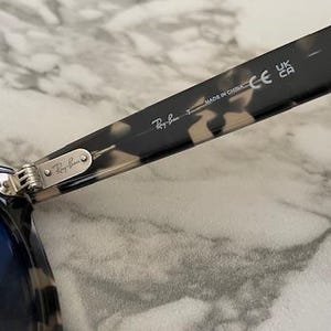 Puede incluir: Primer plano de gafas de sol Ray-Ban con lentes azules. La montura tiene un patr&oacute;n de carey. La bisagra y la patilla de las gafas de sol son visibles, con el logotipo de Ray-Ban y el texto "MADE IN CHINA".