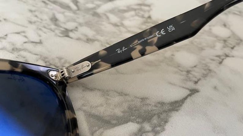 Puede incluir: Primer plano de gafas de sol Ray-Ban con lentes azules. La montura tiene un patr&oacute;n de carey. La bisagra y la patilla de las gafas de sol son visibles, con el logotipo de Ray-Ban y el texto "MADE IN CHINA".