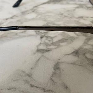 May include: Close-up of eyeglasses with a dark metal frame and black temple tips. The glasses have a sleek, modern design, with a thin frame and a subtle, curved shape. The lenses are not visible in this side view.