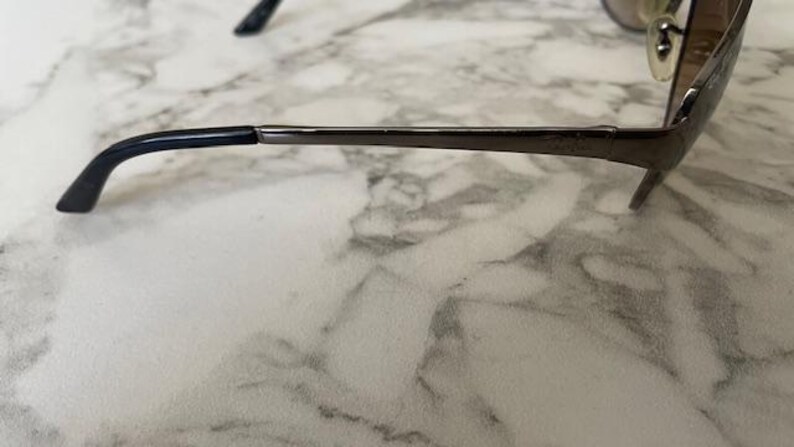May include: Close-up of eyeglasses with a dark metal frame and black temple tips. The glasses have a sleek, modern design, with a thin frame and a subtle, curved shape. The lenses are not visible in this side view.