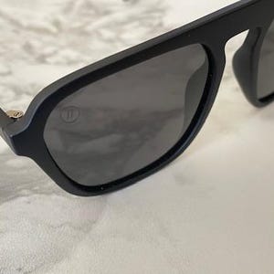 Puede incluir: Gafas de sol negro mate con un dise&ntilde;o de montura de parte superior plana y lentes gris oscuro. Un peque&ntilde;o logotipo circular est&aacute; grabado en la esquina superior de la lente izquierda y un detalle de bisagra plateado es visible en la patilla.