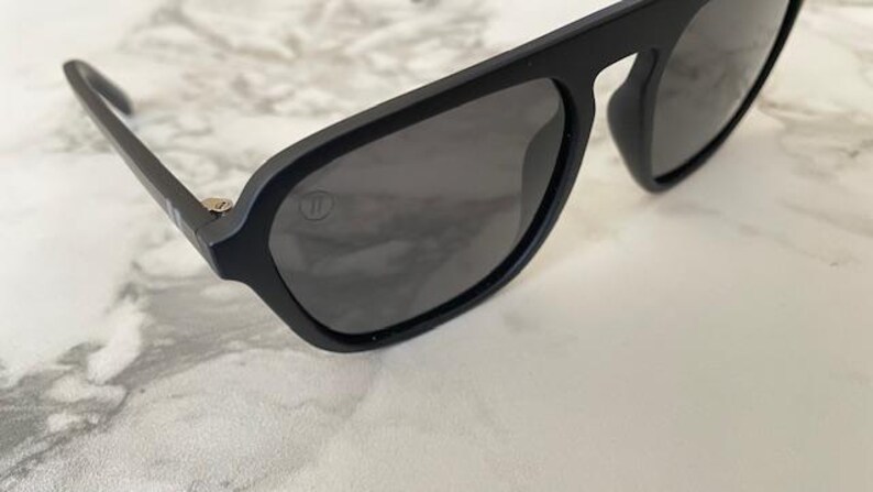 Puede incluir: Gafas de sol negro mate con un dise&ntilde;o de montura de parte superior plana y lentes gris oscuro. Un peque&ntilde;o logotipo circular est&aacute; grabado en la esquina superior de la lente izquierda y un detalle de bisagra plateado es visible en la patilla.