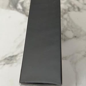 K&ouml;nnte beinhalten: Gezeigt wird eine rechteckige, schwarze Schachtel, vermutlich f&uuml;r Verpackungszwecke. Die Schachtel ist in klarem Kunststoff verpackt, und die Oberseite und die Seiten sind einfarbig matt schwarz. Die Schachtel steht vertikal auf einer hellen, marmorierten Oberfl&auml;che.