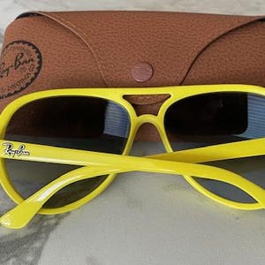 May include: Bright yellow sunglasses with dark lenses and the Ray-Ban logo on the temples. The sunglasses are paired with a brown leather case. The sunglasses have a classic aviator style.