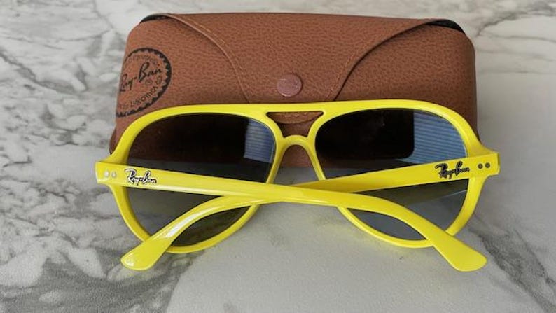 May include: Bright yellow sunglasses with dark lenses and the Ray-Ban logo on the temples. The sunglasses are paired with a brown leather case. The sunglasses have a classic aviator style.
