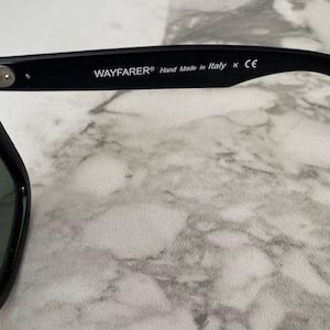 K&ouml;nnte beinhalten: Schwarze Wayfarer-Sonnenbrille mit gr&uuml;nen Gl&auml;sern. Der rechte B&uuml;gel tr&auml;gt den Text "WAYFARER&reg; Hand Made in Italy K CE". Die Sonnenbrille liegt auf einem Marmorhintergrund.
