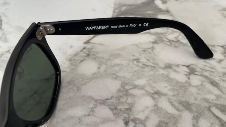 K&ouml;nnte beinhalten: Schwarze Wayfarer-Sonnenbrille mit gr&uuml;nen Gl&auml;sern. Der rechte B&uuml;gel tr&auml;gt den Text "WAYFARER&reg; Hand Made in Italy K CE". Die Sonnenbrille liegt auf einem Marmorhintergrund.