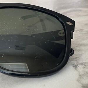 Puede incluir: Gafas de sol negras con lentes oscuras. Las gafas tienen un dise&ntilde;o de montura rectangular cl&aacute;sico. Las lentes son ligeramente reflectantes y la montura tiene un peque&ntilde;o acento plateado en las patillas. Las gafas descansan sobre una superficie de m&aacute;rmol.
