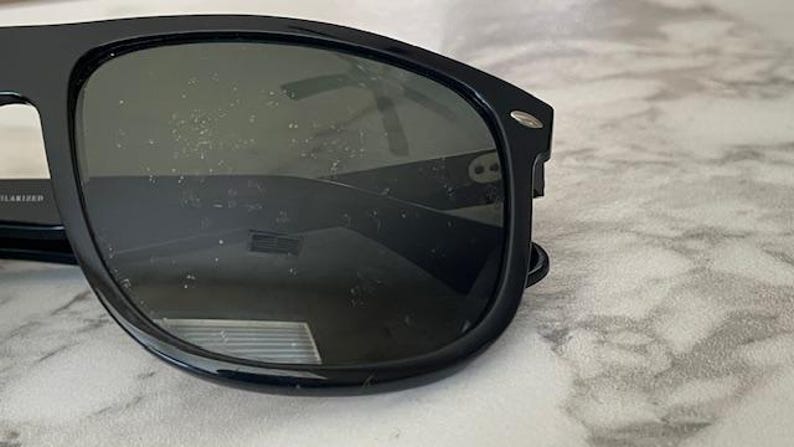 Puede incluir: Gafas de sol negras con lentes oscuras. Las gafas tienen un dise&ntilde;o de montura rectangular cl&aacute;sico. Las lentes son ligeramente reflectantes y la montura tiene un peque&ntilde;o acento plateado en las patillas. Las gafas descansan sobre una superficie de m&aacute;rmol.