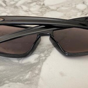 May include: Black sunglasses with dark lenses, folded. The sunglasses have a rectangular frame and the Oakley logo is visible on the temples. The lenses are a dark brown color.
