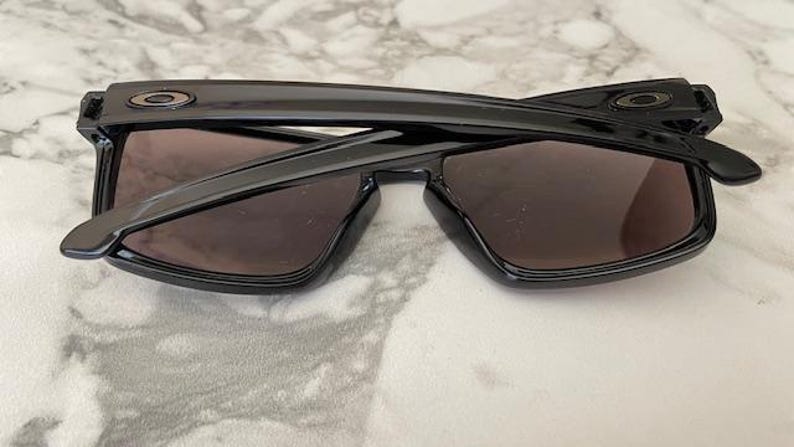 May include: Black sunglasses with dark lenses, folded. The sunglasses have a rectangular frame and the Oakley logo is visible on the temples. The lenses are a dark brown color.