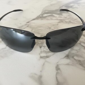 May include: Rimless sunglasses with dark grey lenses and black temples. The sunglasses are set against a white and grey marble background. The lenses are a dark grey colour and have a slight curve.