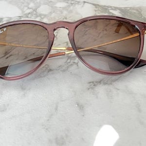May include: Round-framed sunglasses with a translucent mauve frame and brown-tinted lenses. The sunglasses have gold-colored metal arms and the Ray-Ban logo is visible on the left lens. The glasses are resting on a marble surface.