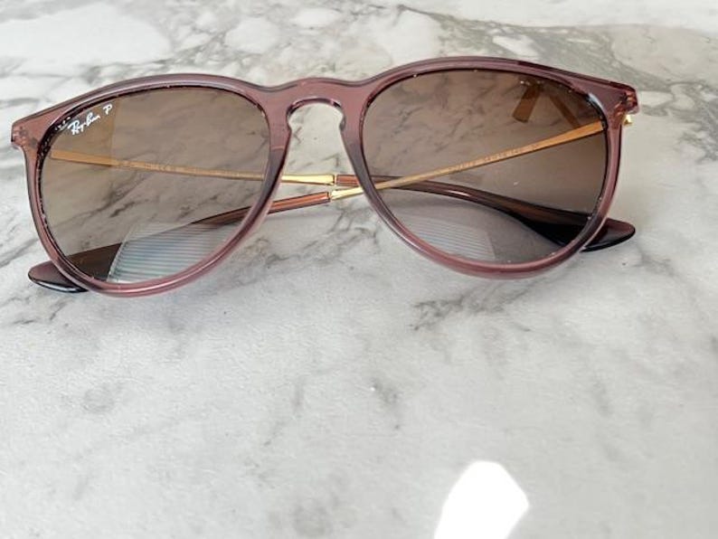 May include: Round-framed sunglasses with a translucent mauve frame and brown-tinted lenses. The sunglasses have gold-colored metal arms and the Ray-Ban logo is visible on the left lens. The glasses are resting on a marble surface.