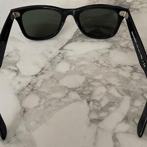 K&ouml;nnte beinhalten: Schwarze Sonnenbrille mit dunklen Gl&auml;sern. Die Sonnenbrille hat ein klassisches, rechteckiges Rahmendesign. Die B&uuml;gel der Sonnenbrille sind gebogen und erstrecken sich nach au&szlig;en. Die Sonnenbrille liegt auf einem Marmorhintergrund.