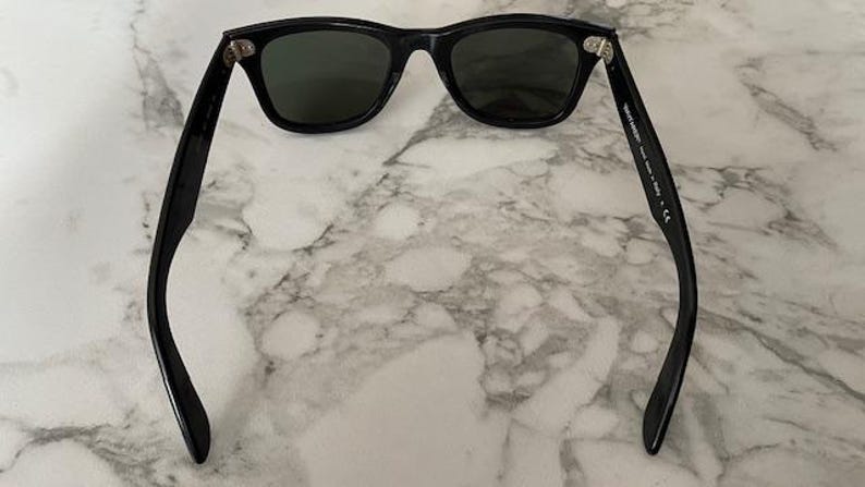 K&ouml;nnte beinhalten: Schwarze Sonnenbrille mit dunklen Gl&auml;sern. Die Sonnenbrille hat ein klassisches, rechteckiges Rahmendesign. Die B&uuml;gel der Sonnenbrille sind gebogen und erstrecken sich nach au&szlig;en. Die Sonnenbrille liegt auf einem Marmorhintergrund.