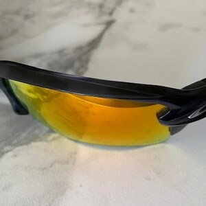 Puede incluir: Gafas de sol deportivas negras con lentes espejadas naranja-amarillo. Las gafas tienen un dise&ntilde;o elegante y curvado con montura negra. Las lentes son de color naranja vibrante, reflejando la luz. Dise&ntilde;adas para deportes y actividades al aire libre.