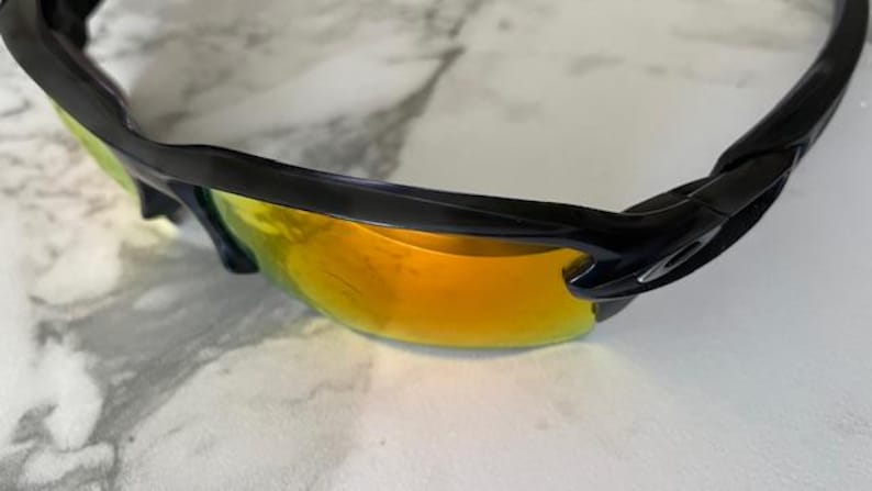 Puede incluir: Gafas de sol deportivas negras con lentes espejadas naranja-amarillo. Las gafas tienen un dise&ntilde;o elegante y curvado con montura negra. Las lentes son de color naranja vibrante, reflejando la luz. Dise&ntilde;adas para deportes y actividades al aire libre.
