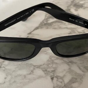 K&ouml;nnte beinhalten: Schwarze Wayfarer-Sonnenbrille mit dunklen Gl&auml;sern. Das Ray-Ban-Logo ist auf dem Rahmen sichtbar. Die Sonnenbrille wird in Italien hergestellt. Die B&uuml;gel sind aufgeklappt.