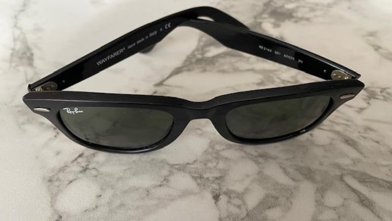 K&ouml;nnte beinhalten: Schwarze Wayfarer-Sonnenbrille mit dunklen Gl&auml;sern. Das Ray-Ban-Logo ist auf dem Rahmen sichtbar. Die Sonnenbrille wird in Italien hergestellt. Die B&uuml;gel sind aufgeklappt.